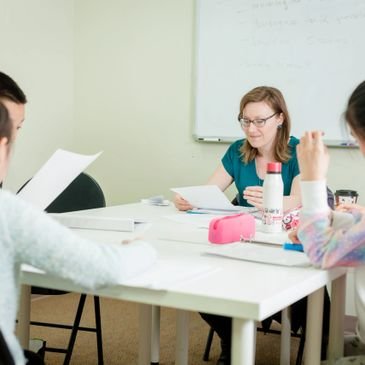 A teacher seated at a table with a small group of students in front of a whiteboard