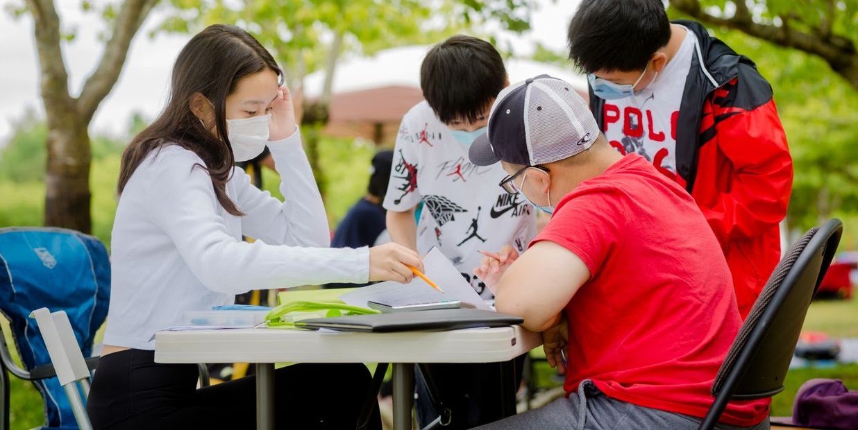 Students and a teacher working together around a table outdoors under the trees